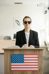 A visually impaired woman reads a Braille book at a podium with an American flag indoors.