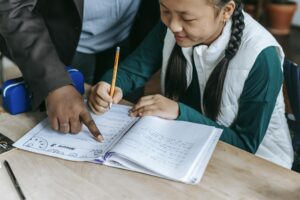 High angle of crop black teacher helping Asian girl with assignment while writing on paper in classroom