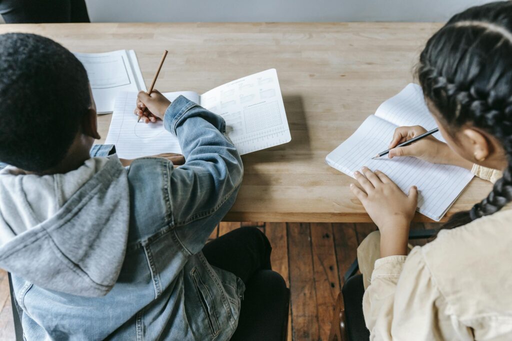 High angle of faceless ethnic schoolchildren in casual clothes sitting at wooden desk with notebooks and pencils while writing task during lesson