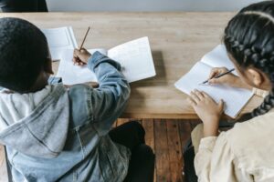 High angle of faceless ethnic schoolchildren in casual clothes sitting at wooden desk with notebooks and pencils while writing task during lesson