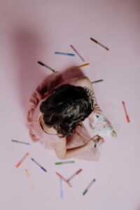 Overhead view of a young girl drawing with markers on a pink background.