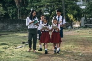 Students in uniforms walking with a teacher outdoors, holding books and papers.